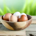 A rustic wooden bowl filled with fresh chicken eggs, both white and brown shells, on a wooden table. The background features softly blurred green plan